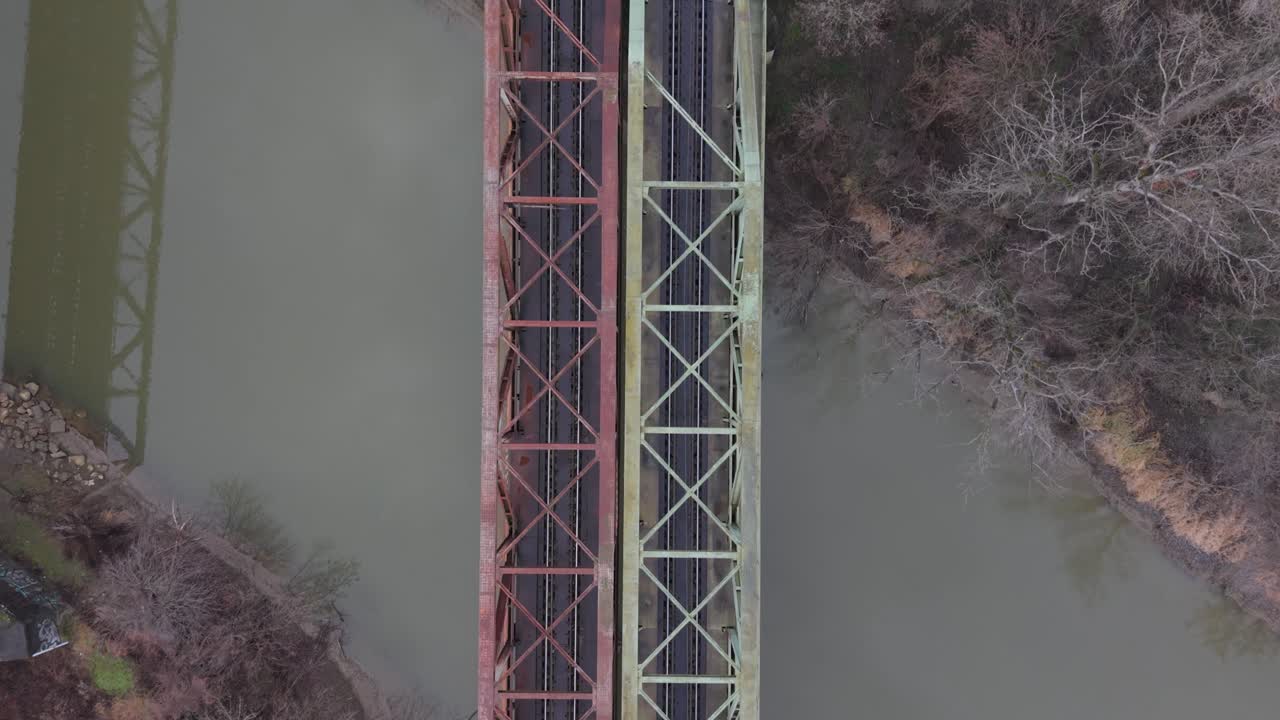 Una veduta aerea dall'alto di ponti ferroviari paralleli che attraversano un fiume calmo. Le strutture in acciaio simmetriche e la vegetazione invernale circostante enfatizzano l'infrastruttura di trasporto e il design industriale.