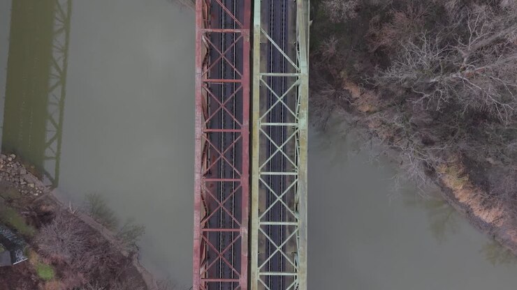 A top-down aerial view of parallel railway bridges crossing a calm river. Symmetrical steel structures and surrounding winter vegetation emphasize transportation infrastructure and industrial design