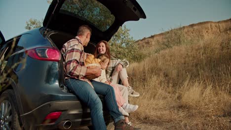 A-happy-family-sits-in-the-trunk-of-an-open-black-car-during-their-picnic-and-relaxation-outside-the-city,-with-a-lot-of-dry-grass-and-small-trees-around.-A-happy-brunette-man-with-a-little-gray-hair-talks-at-a-picnic-with-his-wife,-a-brunette-girl-in-a-green-checkered-shirt,-and-a-little-daughter,-