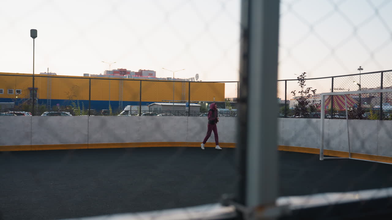 Female fitness instructor runs along curved outdoor court beneath twilight sky, seen through chain link fence, pastel sunset glow lighting yellow industrial building and high rise apartments at dusk