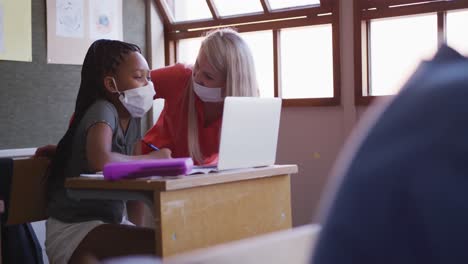 maestra y niña con máscara facial usando una computadora portátil en clase en la escuela