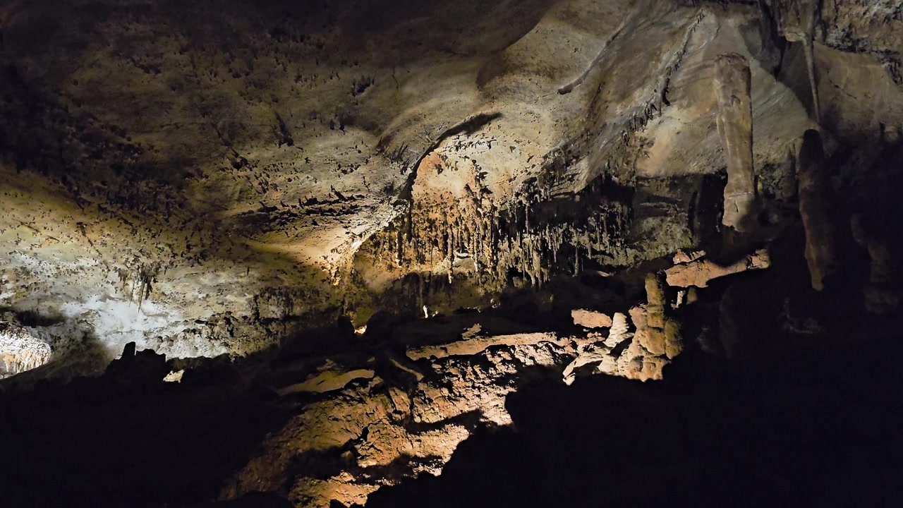 Ancient limestone cavern with rough rock floor, stalagmites rising from the ground, stalactites overhead, cool cave lighting, dark shadows, and scenic underground formations.