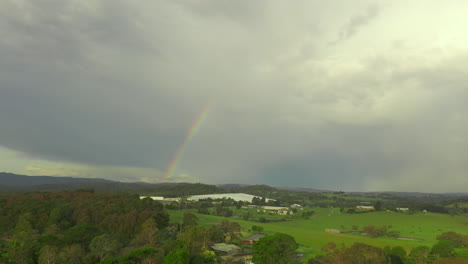 Die-Luftaufnahme-Schwenkt-Nach-Rechts-Und-Zeigt-Gewächshäuser-In-Einer-Natürlichen-Landschaft-Unter-Einem-Regenbogen-Mit-Schweren-Wolken-Am-Himmel