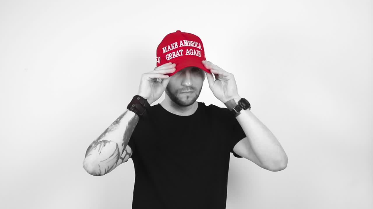 Grayscale footage of a tattooed short haired and bearded man putting on a red vibrant colored MAGA or Make America Great Again Trump presidential hat. Man crosses his arms and nods at the camera happy