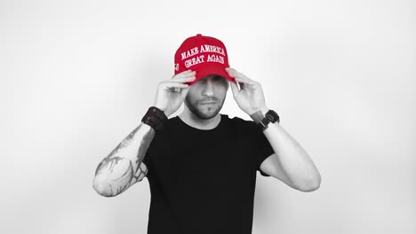 grayscale footage of a tattooed short haired and bearded man putting on a red vibrant colored maga or make america great again trump presidential hat. man crosses his arms and nods at the camera happy