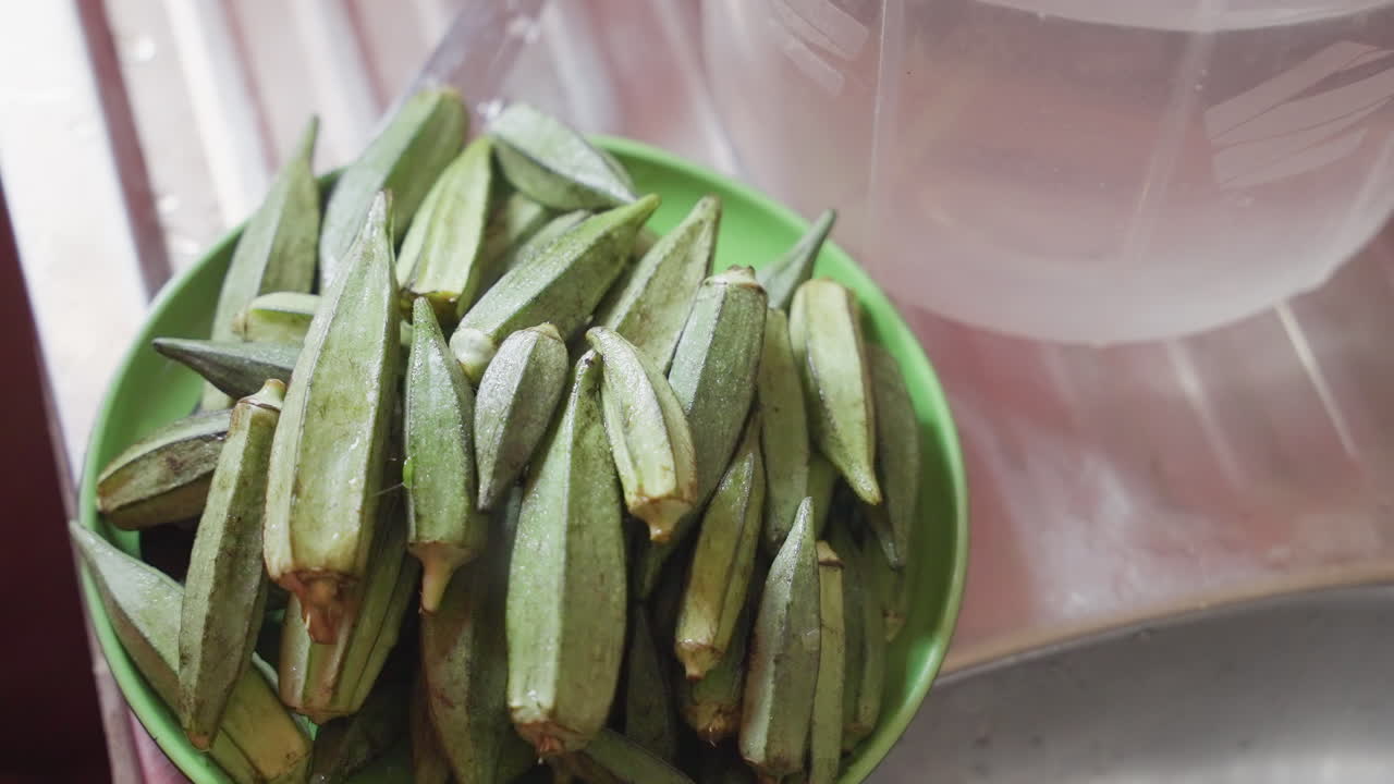 Mangkuk okra segar di meja dapur, juru masak rumahan menata tumpukan, penjual pasar memamerkan hasil bumi dan koki memilih polong untuk resep, tetesan air menempel, sinar matahari menyoroti tekstur bergaris, polong hijau