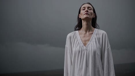 woman in a white dress stands in the rain with her eyes closed, embracing the tranquility of the moment with a peaceful and serene expression on her face