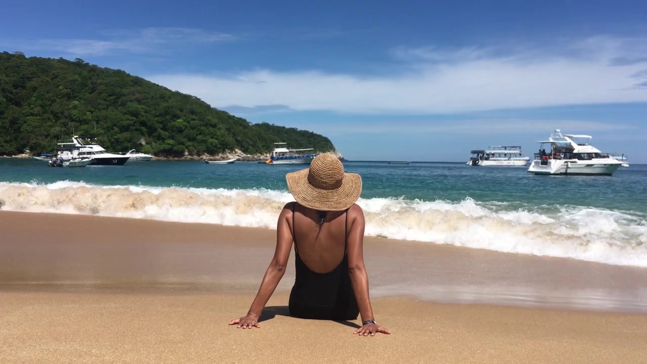 Mulher sentada na praia, apreciando a areia e a água, enquanto observa as águas tropicais e os barcos