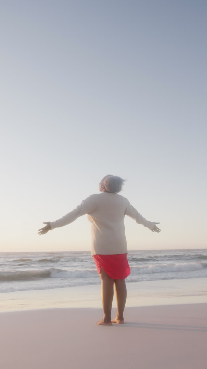 Vertical video of happy senior african american woman widening arms at beach, in slow motion