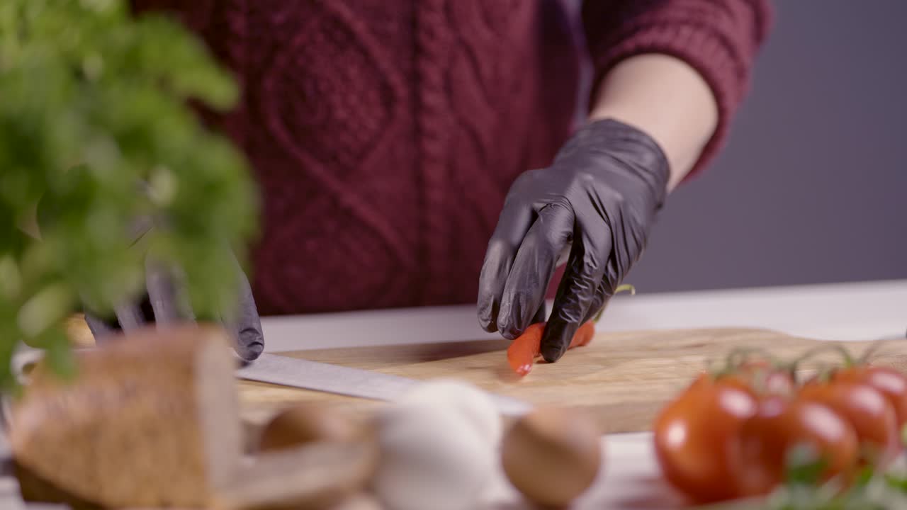 Slow-motion close-up of a chef holding a red chili pepper on a wooden board, preparing to slice it with a sharp knife. Black gloves ensure hygiene, while fresh tomatoes, herbs, and bread add depth