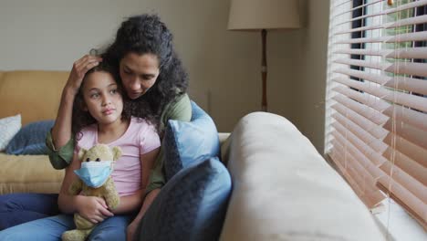 madre e hija de raza mixta abrazándose, hija sosteniendo un oso con máscara facial