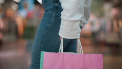 pink shopping bag held by shopper with mint-colored bag slightly visible, set against vibrant mall background featuring colorful reflections, bustling activity