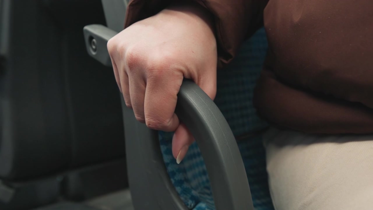 Vista cercana muestra la mano derecha sujetando firmemente el reposabrazos de plástico moldeado en un asiento tapizado con estampado azul, con los dedos curvados, un anillo visible, manga oscura, pantalones beige y una postura sentada tranquila.