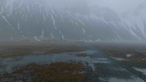 Drone-Shot-of-Misty-Landscape-of-Iceland,-Wetlands-and-Viking-Village-Under-Mountain-Hills