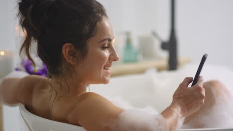 Cheerful-woman-lying-in-bath-with-smartphone