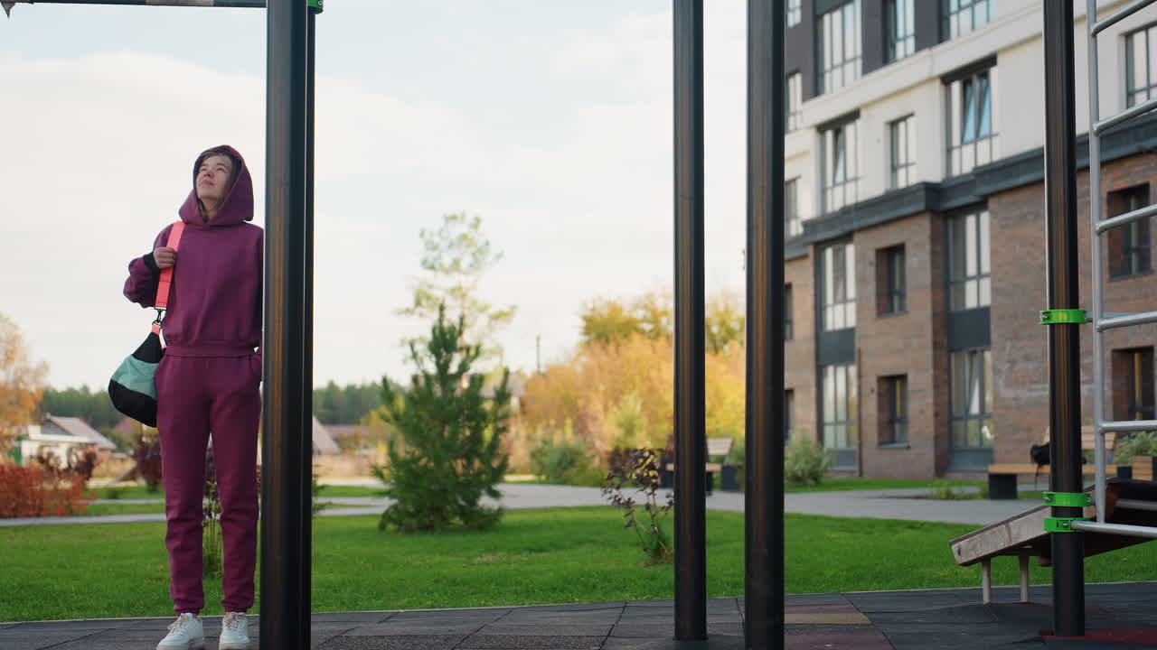 View between vertical bars of workout participant in maroon hoodie carrying bag standing in outdoor gym park looking up at iron structure against autumn urban backdrop with warm lighting