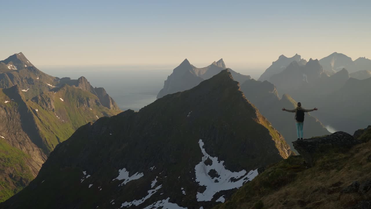Un'escursionista cammina verso uno sperone roccioso e allarga le braccia in segno di libertà, ammirando le mozzafiato montagne e l'oceano delle Lofoten in una morbida luce naturale