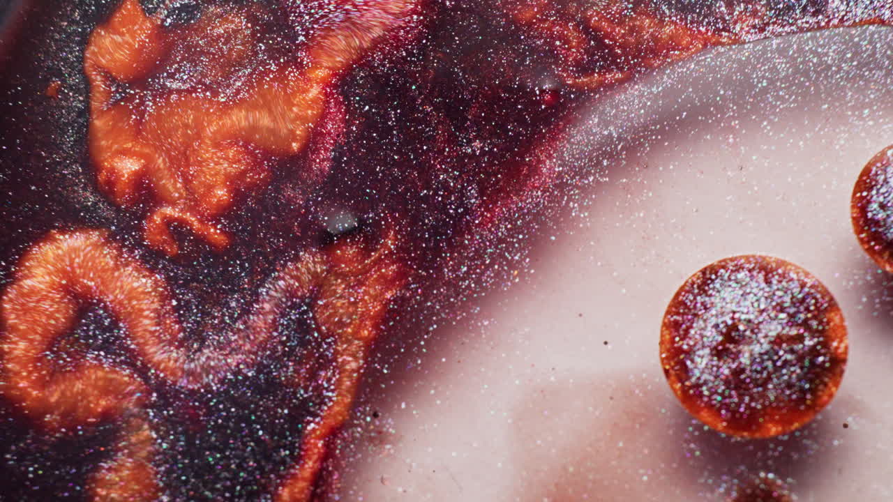 Macro di inchiostro glitterato marrone, arancione e rosso che vortica attorno a una gocciolina poggiata su uno sfondo liquido lattiginoso.