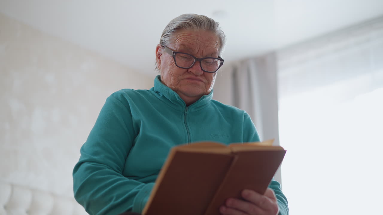 Lonely elderly pensioner in green teal sweater reading a book with focused expression, engaged in her own world of reading. Indoor space with soft lighting creates peaceful, reflective mood