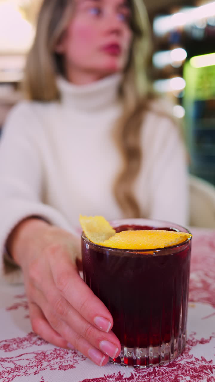 Close up of a woman holding a negroni cocktail on a red and white tablecloth at a restaurant. Vertical