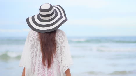 Back-view-Unrecognizable-Woman-Traveler-in-White-Sundress-and-Stripped-Hat-Standing-Seafront-Looking-ar-Sea-Waves-Rolling-Over-Beach---slow-motion