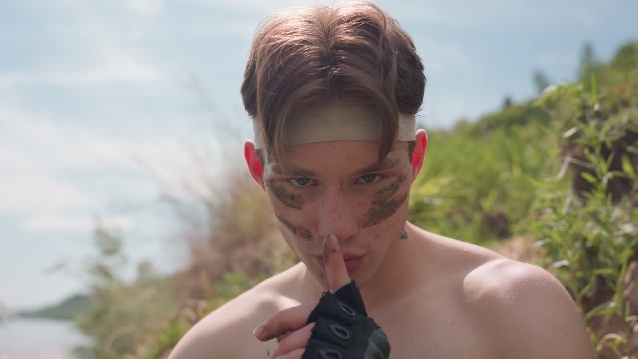 Shirtless fighter with muddy face paint glares intensely under sunlight while raising finger in silence command, exuding intimidation, control, and survival energy amid rugged green hillside terrain