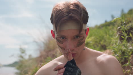 shirtless fighter with muddy face paint glares intensely under sunlight while raising finger in silence command, exuding intimidation, control, and survival energy amid rugged green hillside terrain