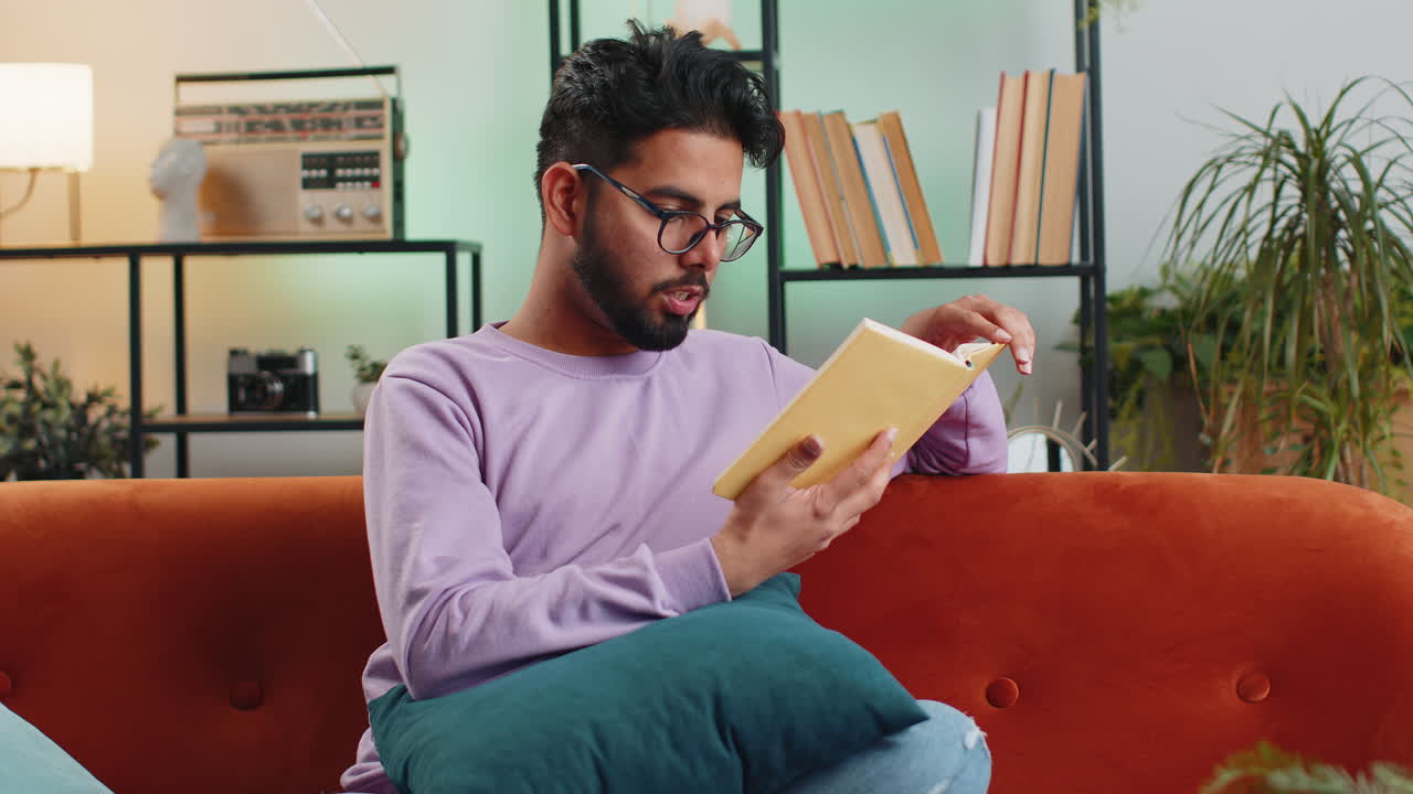 Indian man reading interesting book turning pages smiling enjoying literature taking a rest on sofa