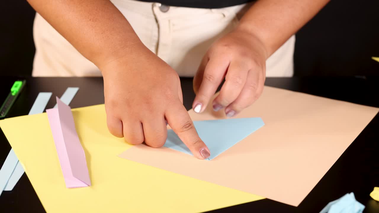 Person folds blue origami paper on yellow sheet, close-up, under bright, even lighting