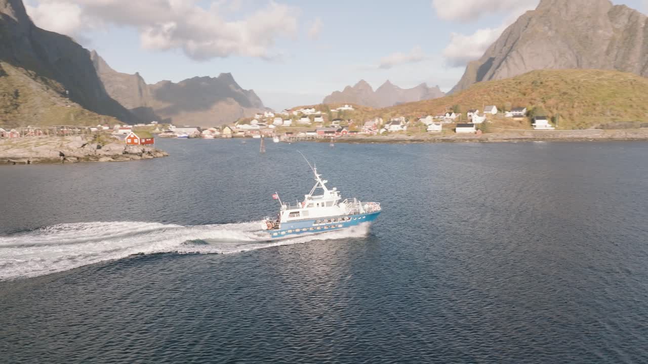 Ett vit- och blåfärgat fiskefartyg far fram genom lugna fjordvatten och skapar dramatiska svallvågor. Höga klippiga berg ramar in en norsk kustby med traditionella röda hus. Gyllene timmen.