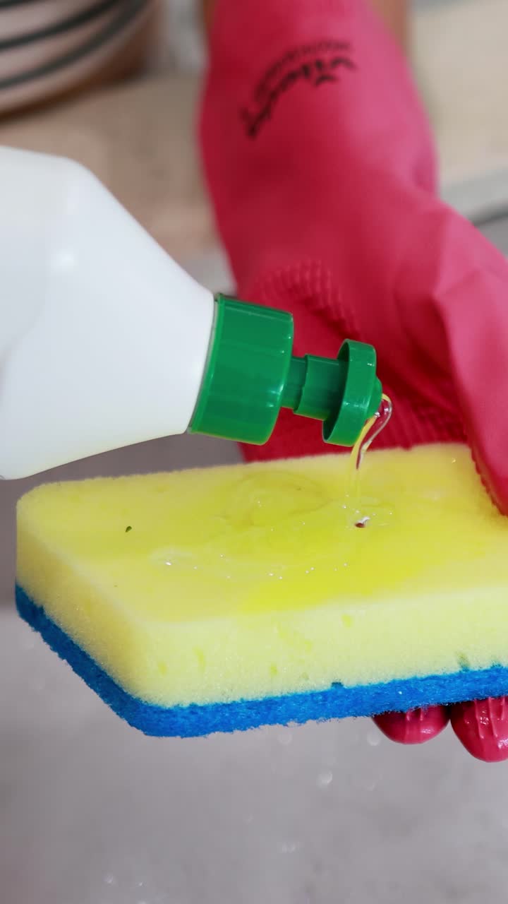 A person wearing pink gloves pours dish soap onto a yellow sponge in a kitchen environment