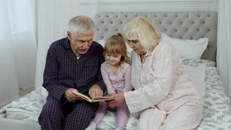 cute girl with senior retired grandmother and grandfather sitting on bed reading book in bedroom