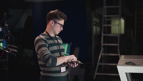 man wearing grey striped sweater and glasses adjusts wireless audio transmitter on video production set indoors, with ladder, monitor and gear in dimly lit studio background, focusing on sound setup