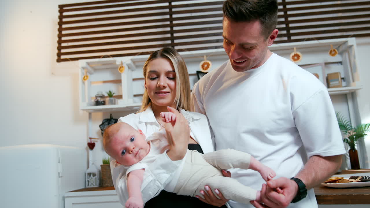 Beautiful blonde woman holding her newborn baby indoors. Husband and happy dad stands beside touching child's bare feet.