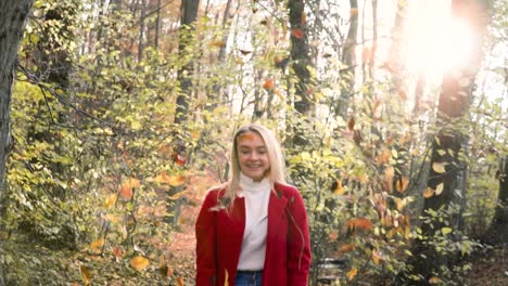 slowmotion young beautiful woman throwing leaves in the air amidst thee orange brown autumn forest woodland while wearing a red coat