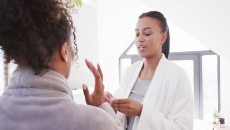 feliz pareja de lesbianas biraciales en albornoces aplicando máscaras debajo de los ojos en un baño soleado, cámara lenta