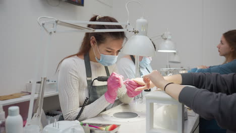 nail technicians wearing protective masks and pink gloves performing detailed manicure treatment for two women seated at workstations under bright salon lamps in clean modern nail salon interior