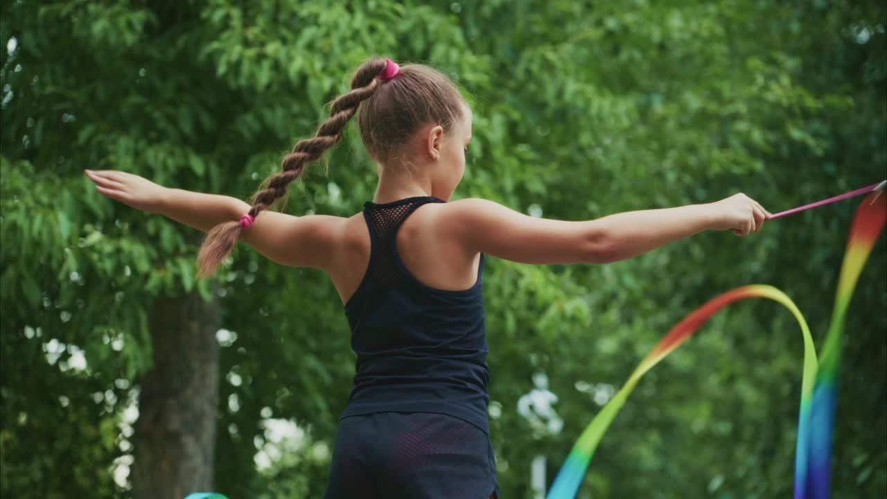A young girl showcases her rhythmic gymnastics skills in a lush green setting, gracefully manipulating colorful ribbons with elegance and precision as she performs
