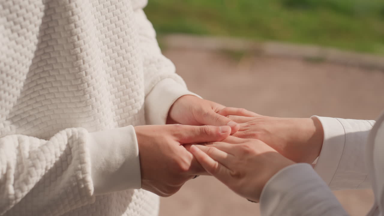 Close hands exchanging wedding ring outdoors, white women exchanging vow with tender touch, sweater texture, sunlight glint on bracelet, intimate family marriage moment, quiet emotion
