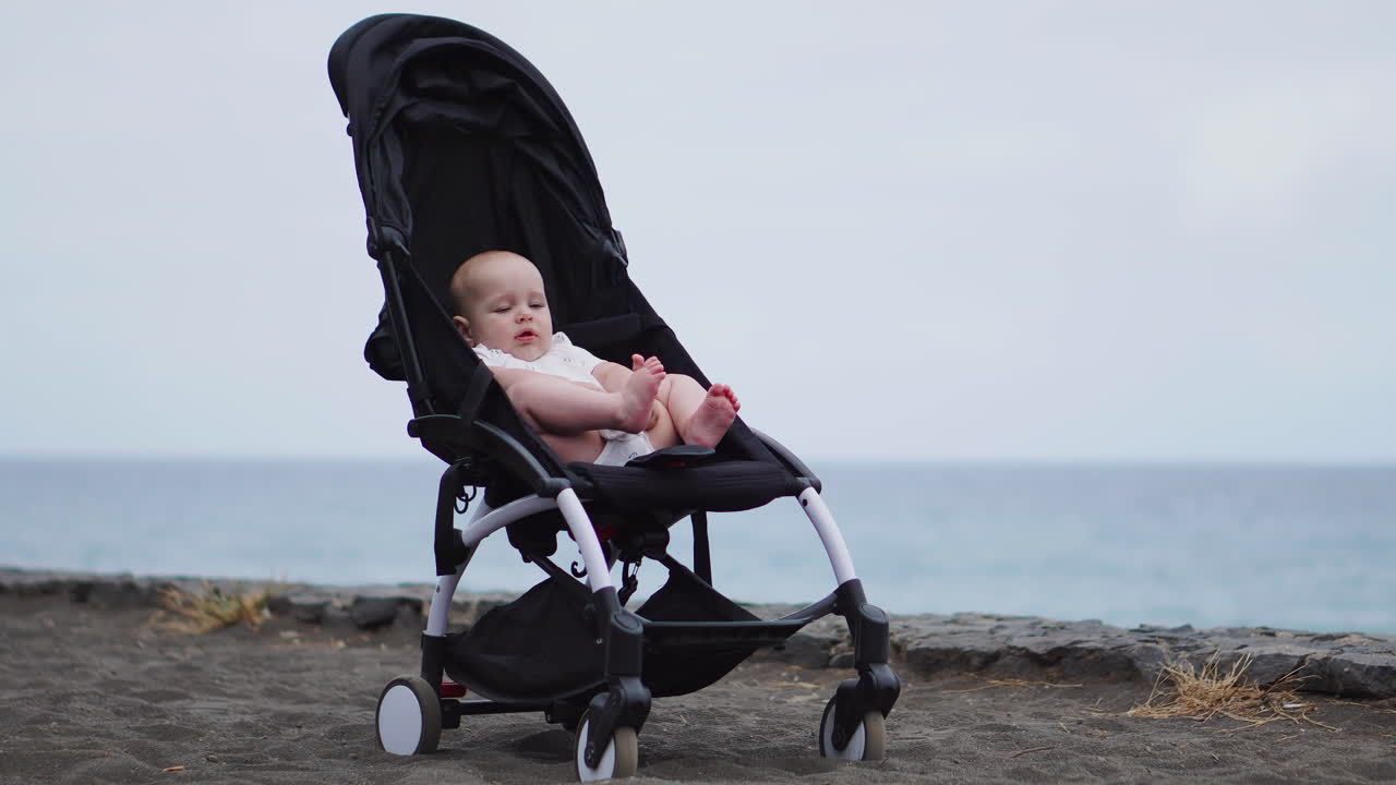 Baby in a stroller on a sandy beach, peacefully looking into infinity and sea waves with great interest, enjoying late summer evening by the sea. Family vacation, traveling with children concept.