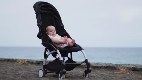 Baby-in-a-stroller-on-a-sandy-beach,-peacefully-looking-into-infinity-and-sea-waves-with-great-interest,-enjoying-late-summer-evening-by-the-sea.-Family-vacation,-traveling-with-children-concept.