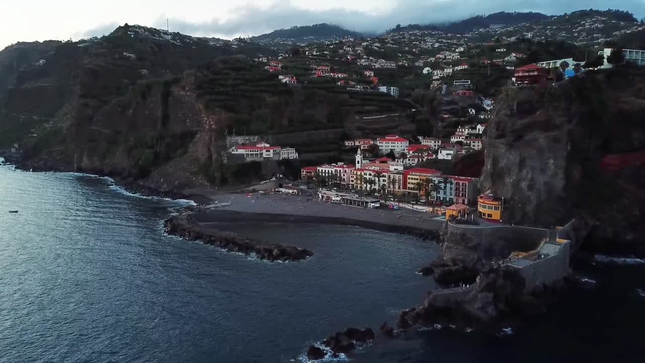 Ponta do Sol, Madeira, at sunset, featuring dark sand beach, colorful houses, volcanic cliffs and the Atlantic ocean crashing against the rocks, slow motion drone footage
