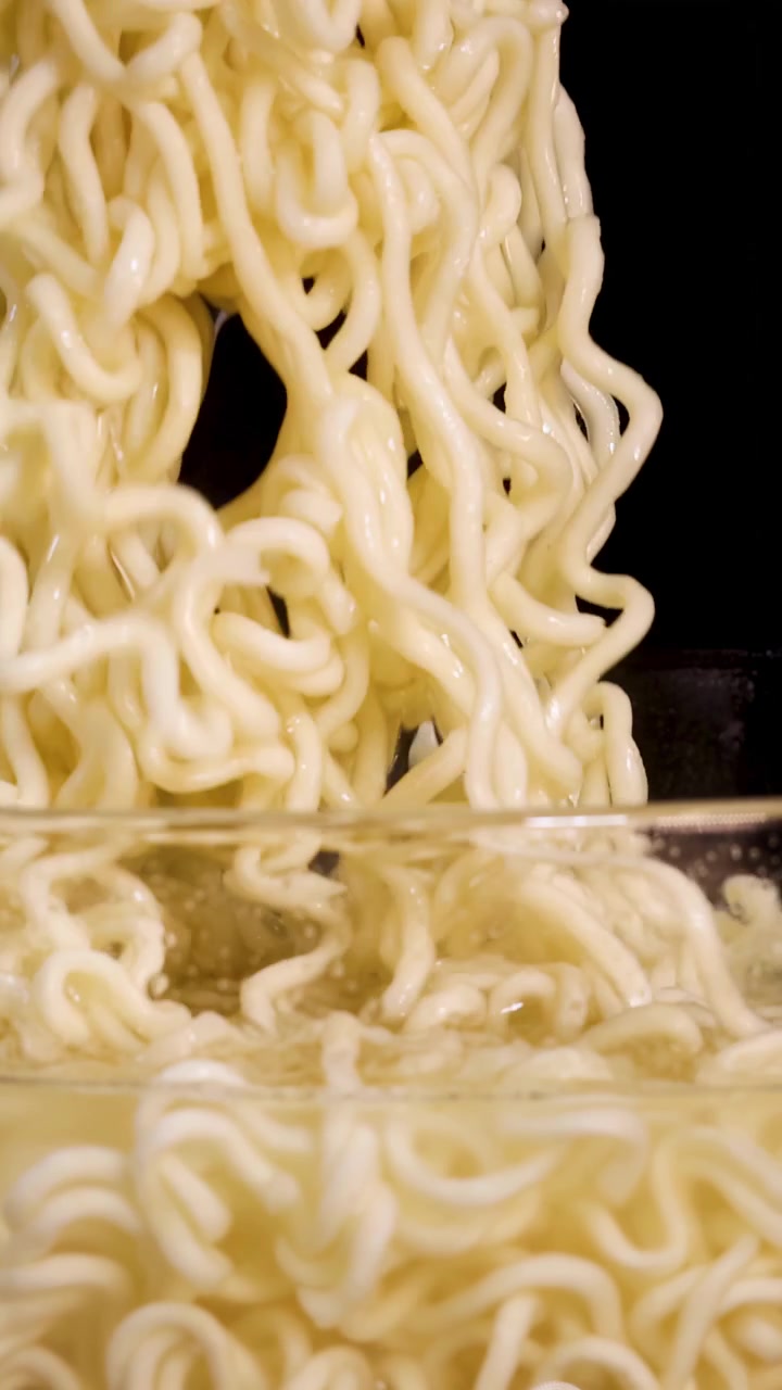 Le bacchette sollevano noodles istantanei cotti da una ciotola di vetro, poi li rilasciano di nuovo nel brodo caldo sotto un'illuminazione drammatica da studio con uno sfondo nero
