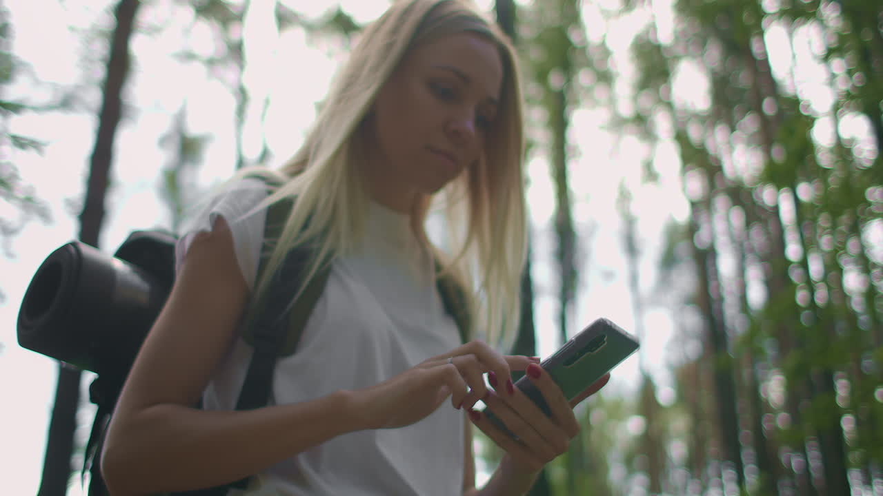 una mujer joven con un teléfono móvil camina por el bosque viajando con una mochila en cámara lenta. navegar por el bosque usando el navegador en su teléfono móvil