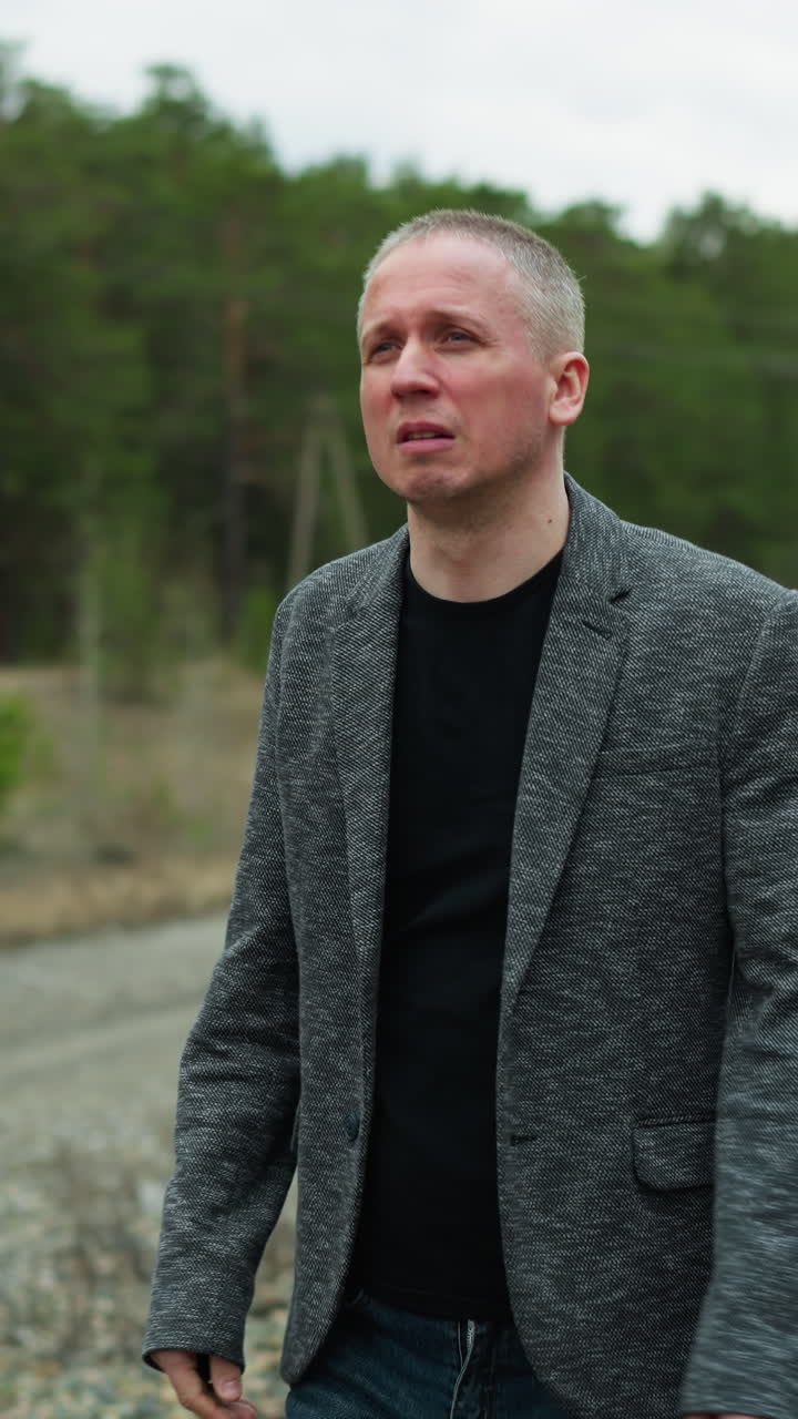 A close-up view of a middle-aged man in a grey suit and black shirt walking along railway tracks in a forested area, the man covers his face with one hand, appearing deep in thought