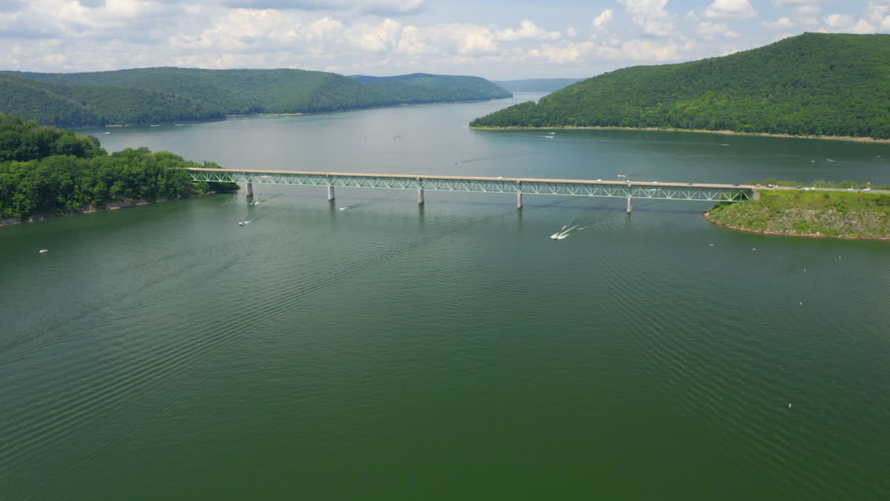 여름철 펜실베니아 숲의 파란색과 녹색 호수 위를 날아가는 공중 드론은 보트와 제트스키가 물 위를 운전하고 자동차가 호수 위의 다리를 건너는 동안
