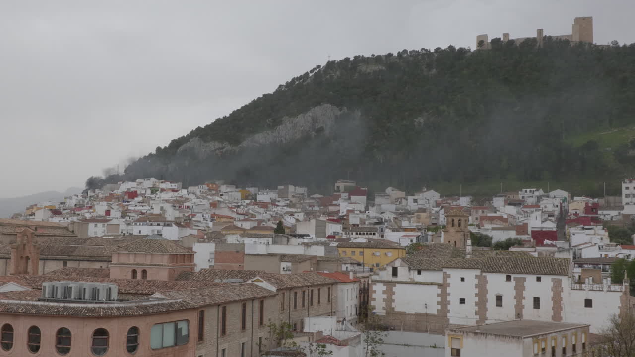 Una gran columna de humo negro de un incendio urbano se eleva sobre los tejados de Jaén, con el Castillo de Santa Catalina al fondo en un día nublado.
