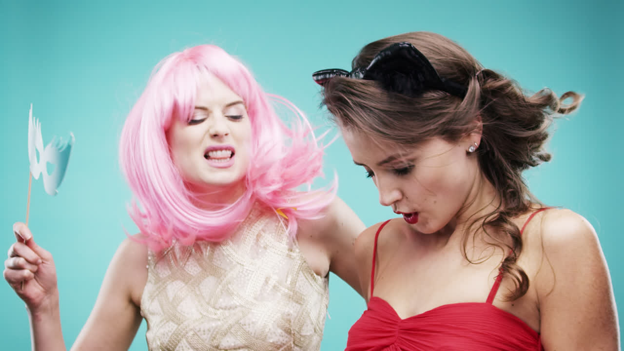 빨간 드레스와 분홍색 머리카락을 입은 여자 친구들이 느린 움직임의 파티 사진 부스에서 춤추고 있습니다.