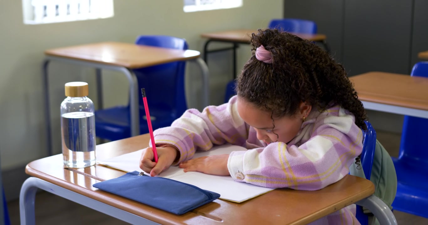 African American schoolgirl writing in notebook with red pencil while doing classwork fidgeting