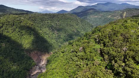 drone camera glides above dense rainforest, revealing a dramatic cliff and river gorge under bright daylight with sweeping, cinematic movement
